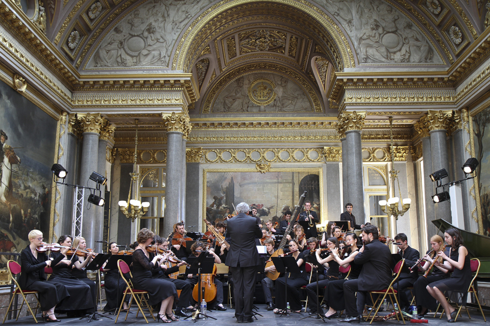 A group of musicians, performing with historical instruments, being conducted by a conductor, seated in an elaborately decorated room in France.
